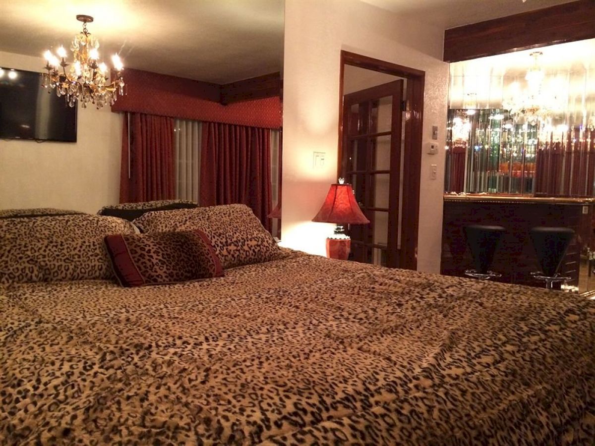 The image shows a bedroom with leopard print bedding, a chandelier, a lamp, a bar area with two stools, and maroon drapes covering a window.