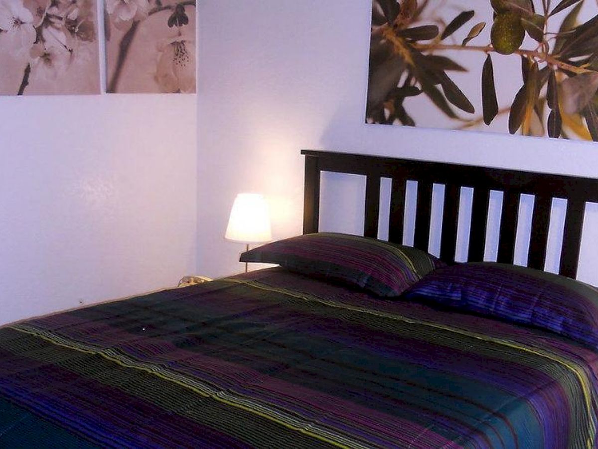 A neatly made bed with dark-colored bedding, a wooden headboard, a table lamp, and floral artwork on the walls are in the room.