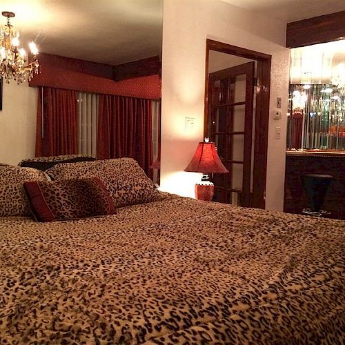 A cozy bedroom with animal print bedding, chandelier, red lamp, bar area with stools, and large window with curtains is shown.