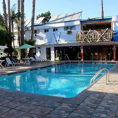 The image shows an outdoor pool surrounded by lounge chairs, umbrellas, and palm trees, with a building offering pizza and sandwiches in the background.