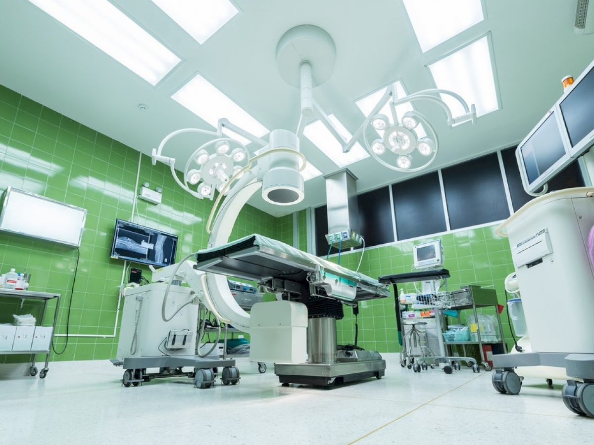 This image shows a modern operating room with green walls, an operating table, medical equipment, monitors, lights, and various surgical tools.