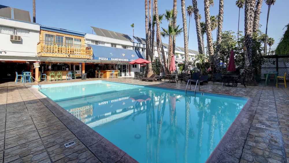 A clear swimming pool surrounded by tall palm trees and buildings with outdoor seating, under a bright blue sky, creating a relaxed atmosphere.