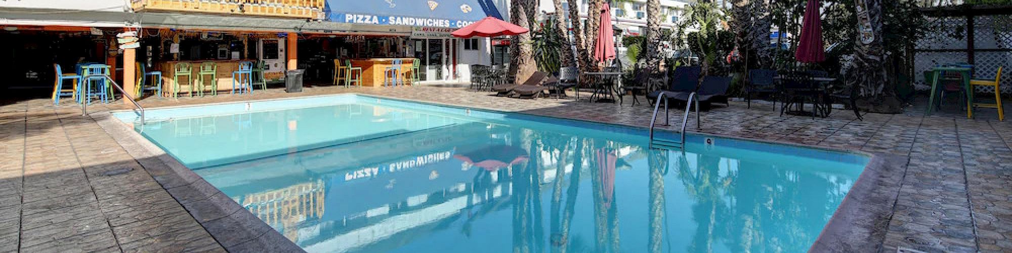 A clear swimming pool surrounded by tall palm trees and buildings with outdoor seating, under a bright blue sky, creating a relaxed atmosphere.