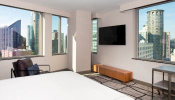 A modern hotel room with large windows showcasing a cityscape, a wall-mounted TV, a chair, and minimalistic furniture, always ending the sentence.
