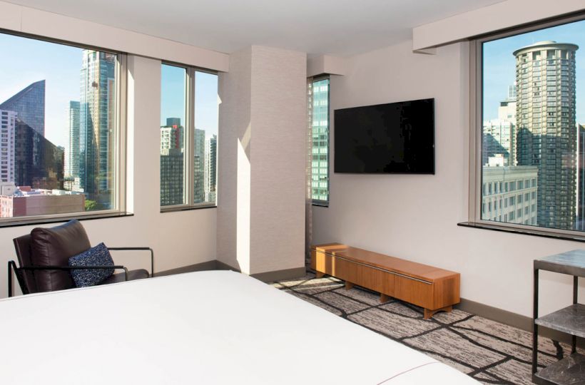 A modern hotel room with large windows showcasing a cityscape, a wall-mounted TV, a chair, and minimalistic furniture, always ending the sentence.