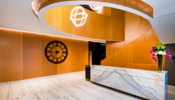 The image shows a modern lobby with a large clock on the wall, wooden accents, a white marble reception desk, and flowers on the counter.