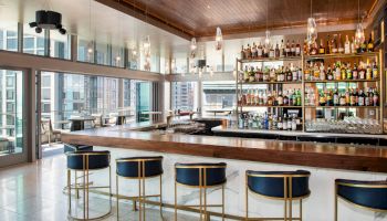 A modern bar with high stools, a wooden counter, and a well-stocked shelf of liquor bottles, surrounded by large windows with a city view.