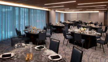 The image shows a modern banquet hall with round tables set with tableware, glassware, and floral centerpieces. The room has dim lighting and blue curtains.