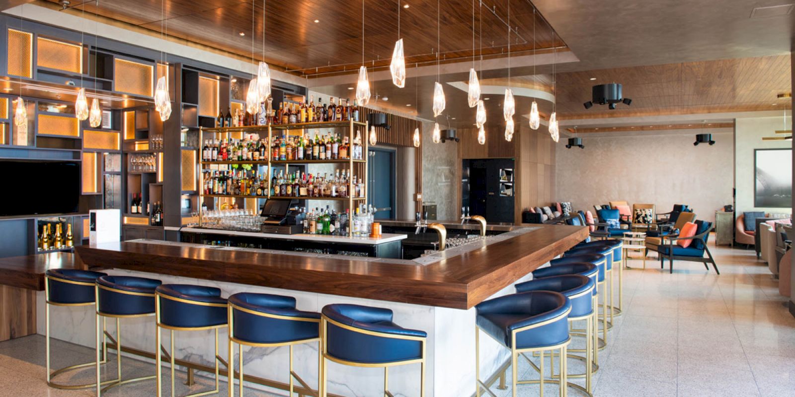 A modern bar with a variety of drinks on shelves, blue bar stools, elegant pendant lights, and a cozy seating area in the background.