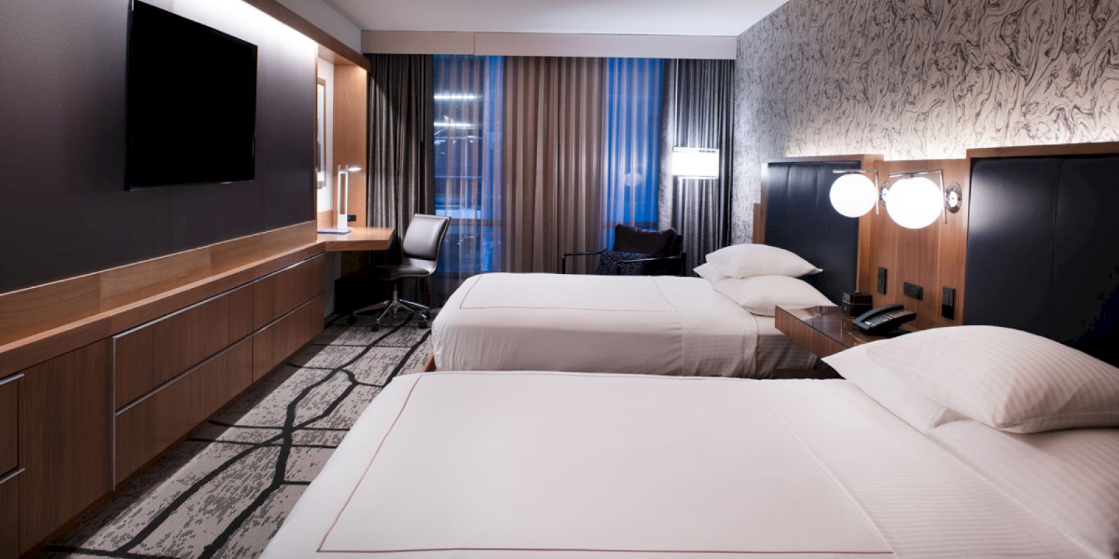 A modern hotel room with two twin beds, a wall-mounted TV, a desk with a chair, and a window with curtains and a view.