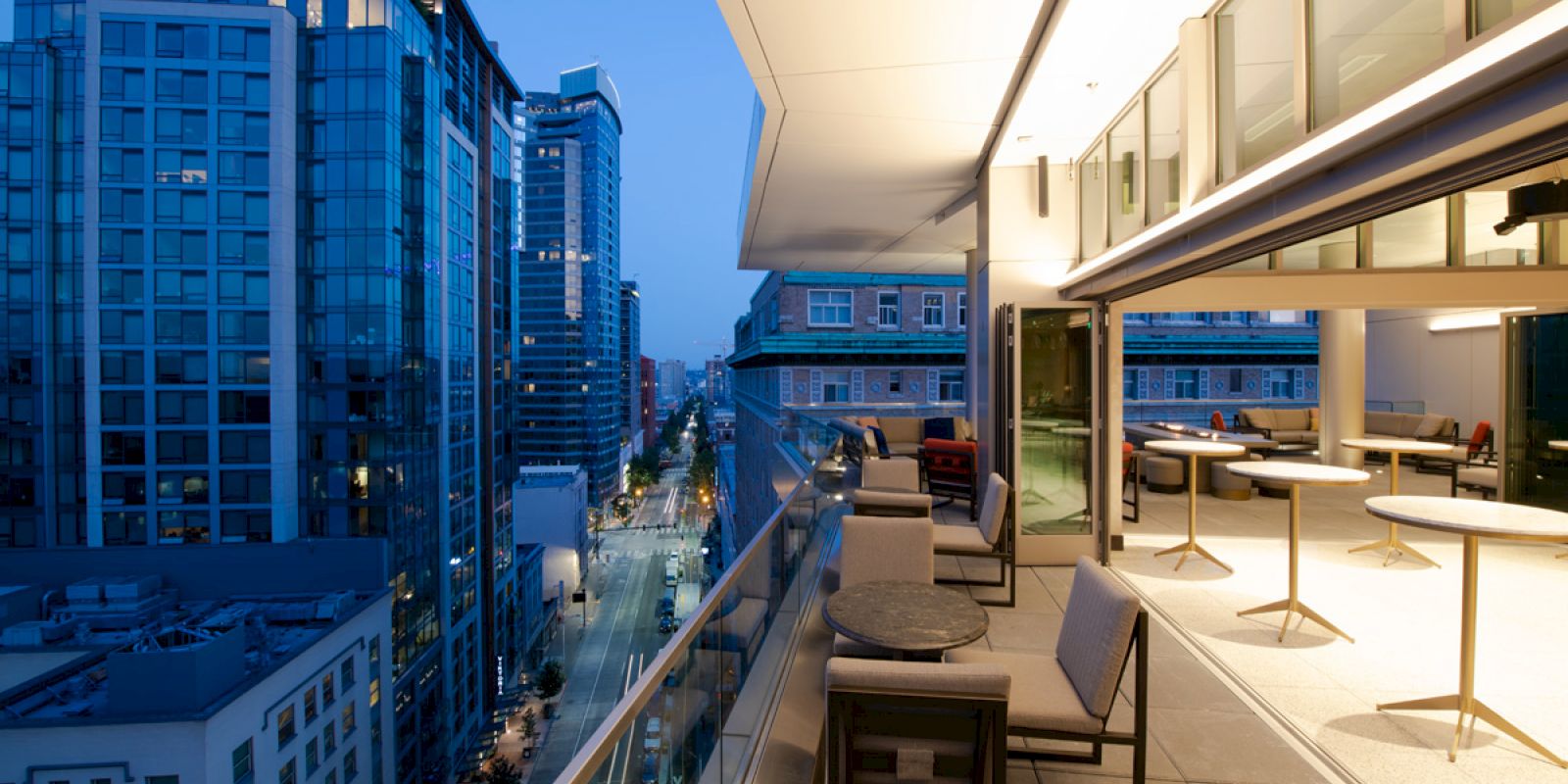 The image shows a modern urban rooftop balcony with seating, overlooking a city street with tall buildings at dusk.