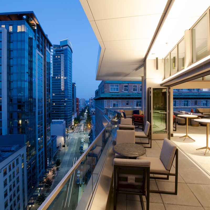 A modern balcony with seating and tables overlooking a city street scene filled with tall buildings during twilight.