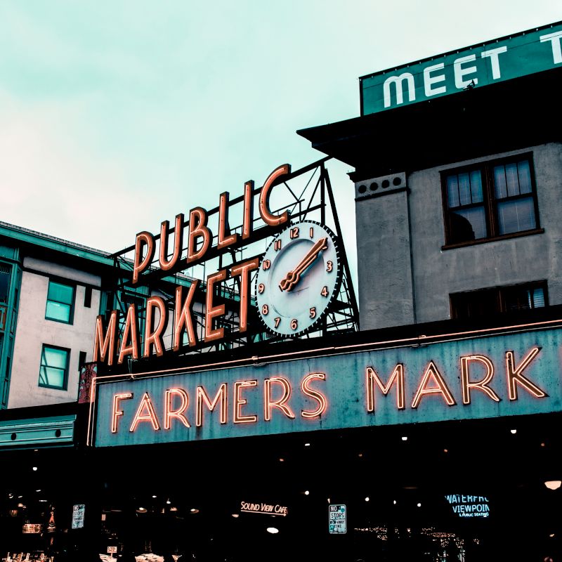 This image shows the neon signs and clock of a public farmers market set amidst city buildings on a cloudy day.