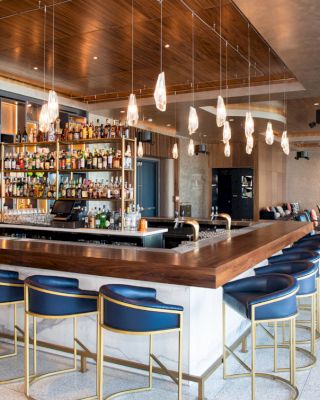 This image shows a modern bar with a wooden counter, blue cushioned stools, hanging lights, and a wall stocked with various bottles in a stylish setting.