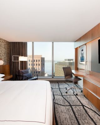 A modern hotel room with a large bed, desk, chair, and television, featuring a city view through expansive windows, and stylish decor.