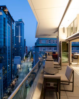 Modern balcony overlooking an urban cityscape at dusk, featuring outdoor seating and tall buildings with lights illuminating the scene.