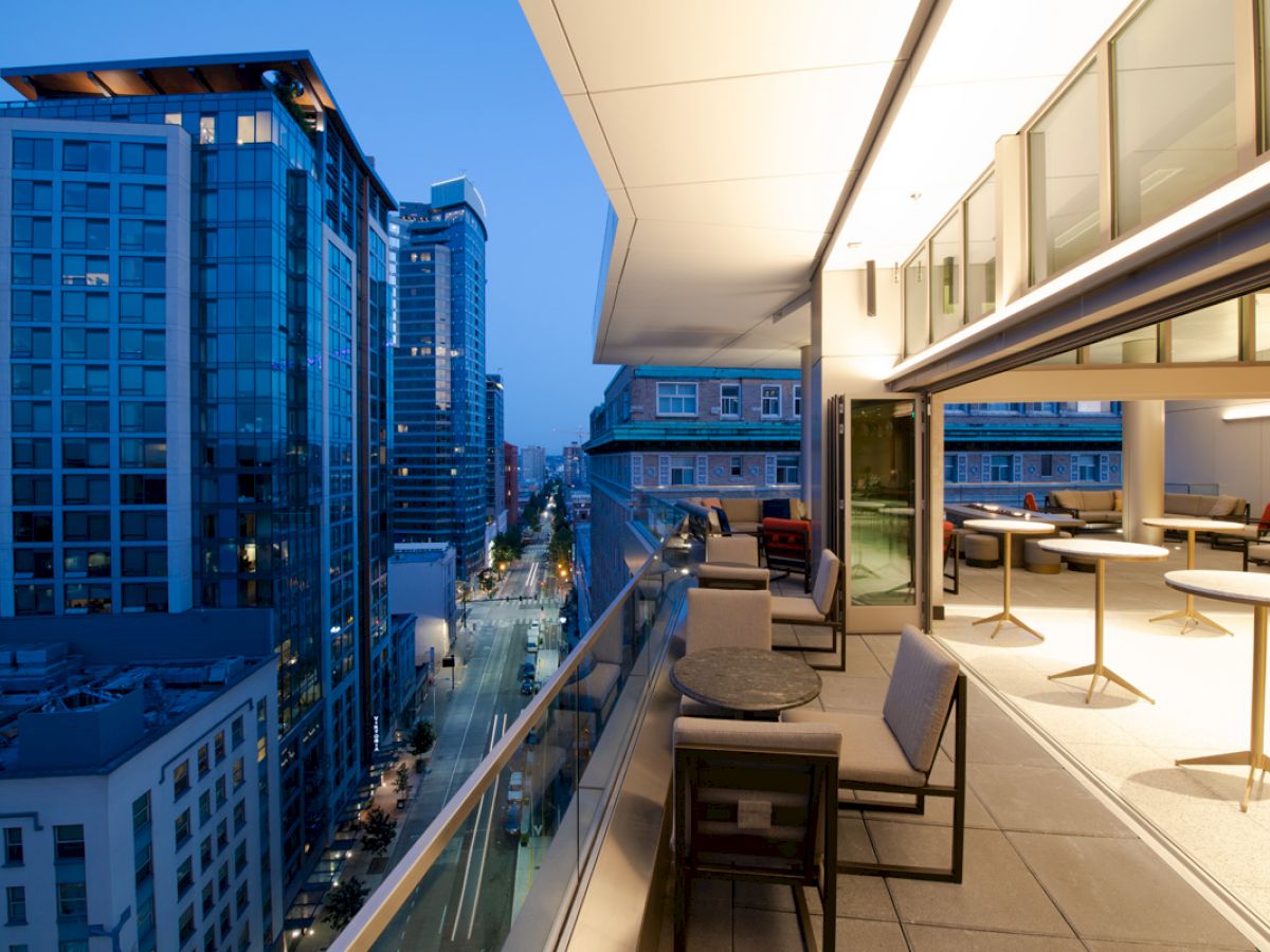 The image shows a modern city balcony at twilight, with stylish outdoor seating, overlooking a street lined with tall buildings.