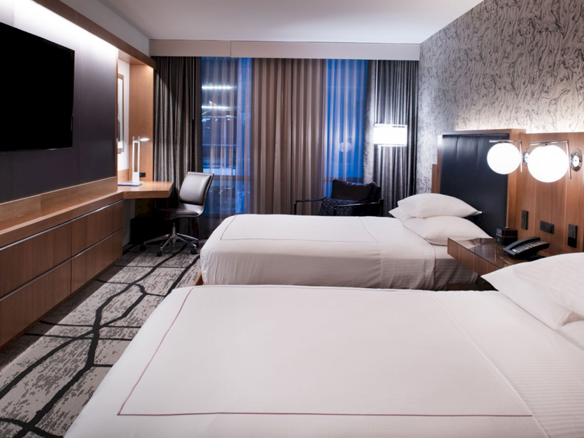 The image shows a modern hotel room with two neatly made beds, a TV on the wall, a desk with a chair, and large windows with curtains.