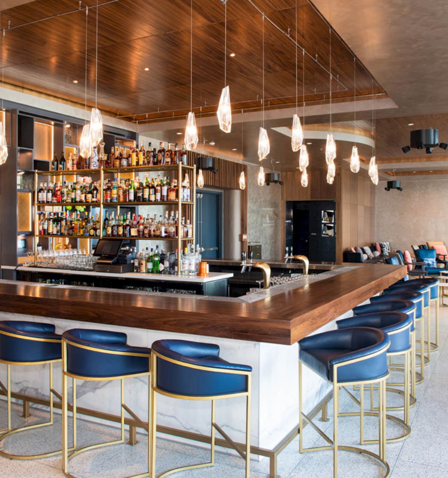 The image shows a modern bar with a wooden counter, blue bar stools, and a well-stocked shelf of liquor bottles behind the counter.