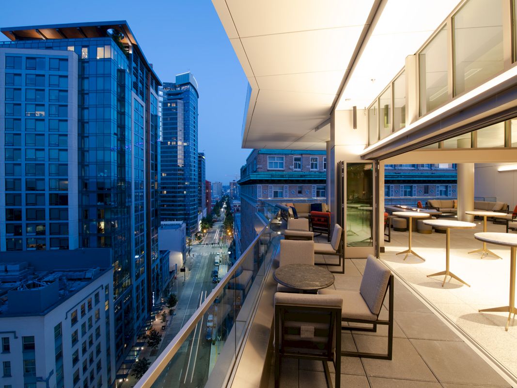 The image showcases a modern urban balcony with outdoor seating overlooking a city street, tall buildings, and city lights during twilight.
