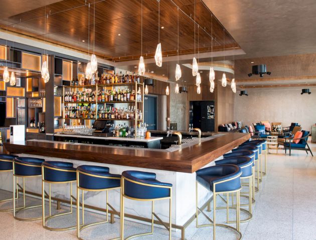 A modern bar with a wooden countertop, blue bar stools, a variety of drinks on shelves behind, stylish lighting, and additional seating areas.