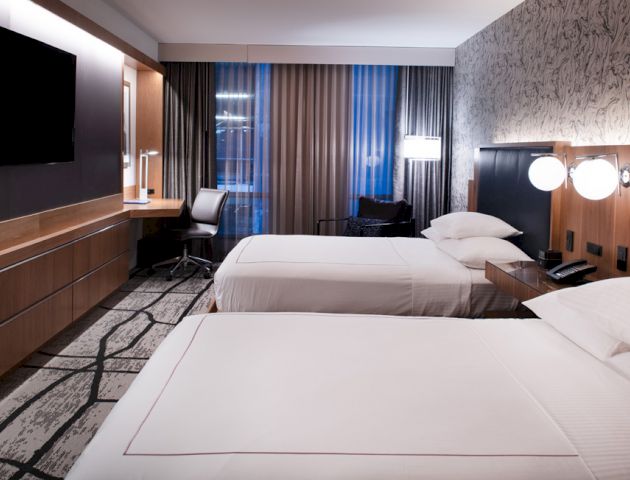 A modern hotel room with two twin beds, a wall-mounted TV, a desk with a chair, and contemporary decor.