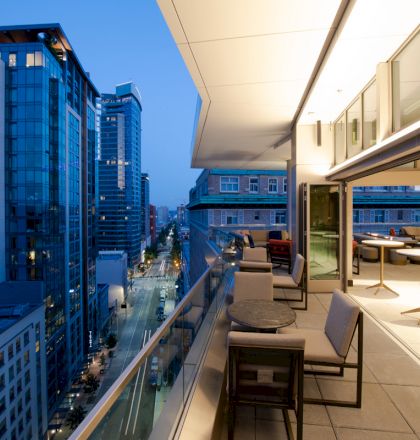 An outdoor balcony area with tables and chairs, overlooking a modern cityscape during twilight. Tall buildings are visible in the background.