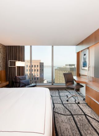 This image shows a modern hotel room with a large bed, a wall-mounted TV, a desk, and a cityscape view through large windows.