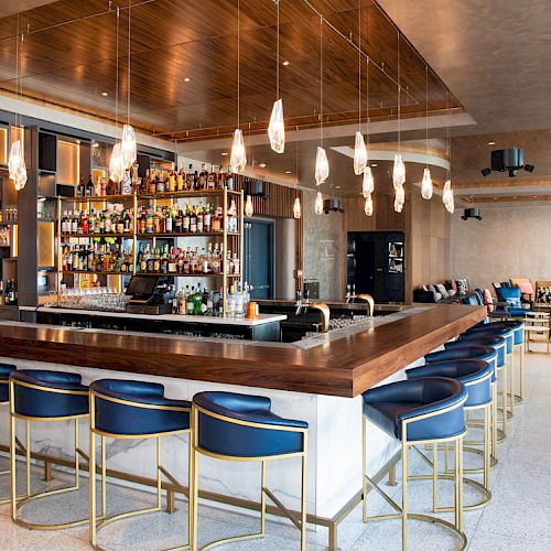 The image shows a modern bar with blue bar stools, a well-stocked central bar, pendant lights, and a seating area in the background.