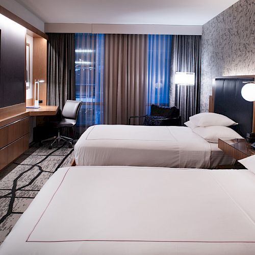 A modern hotel room with two beds, a wall-mounted TV, a desk and chair by the window, and contemporary decor.
