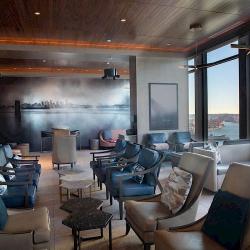 A modern lounge with large windows overlooking a waterfront, featuring comfortable seating, a mural, and a wooden ceiling.