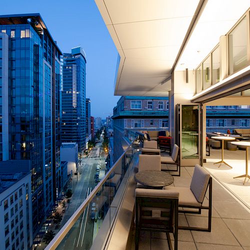 A modern city balcony with several tables and chairs overlooks a street lined with tall buildings at dusk.