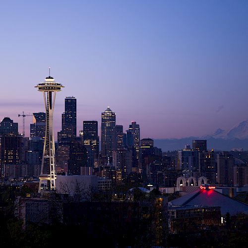 A cityscape at twilight featuring the Seattle skyline, the Space Needle prominently in view, with a distant mountain in the background.