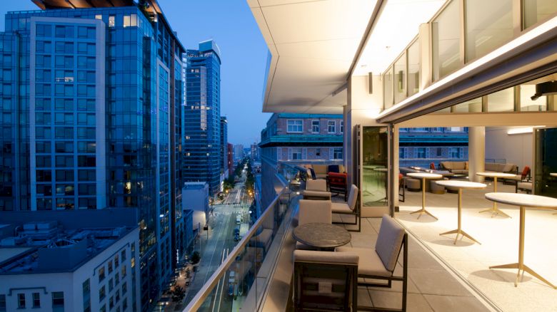 A modern balcony overlooks a cityscape at dusk. The area has seating and tables. Tall buildings line the street in the background.