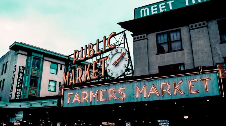 The image shows an iconic sign for a public farmers market with neon lights in an urban setting, featuring a clock and surrounding buildings.