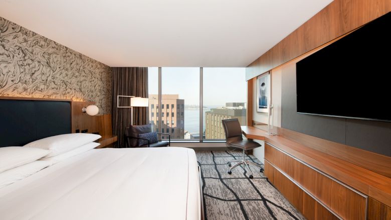 A modern hotel room with a large bed, wall-mounted TV, desk with chair, and a window view of city buildings and water in the distance.