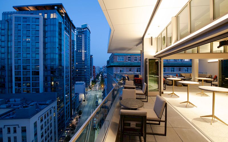 The image shows a modern balcony with tables and chairs, overlooking an urban cityscape with tall buildings and well-lit streets at dusk.