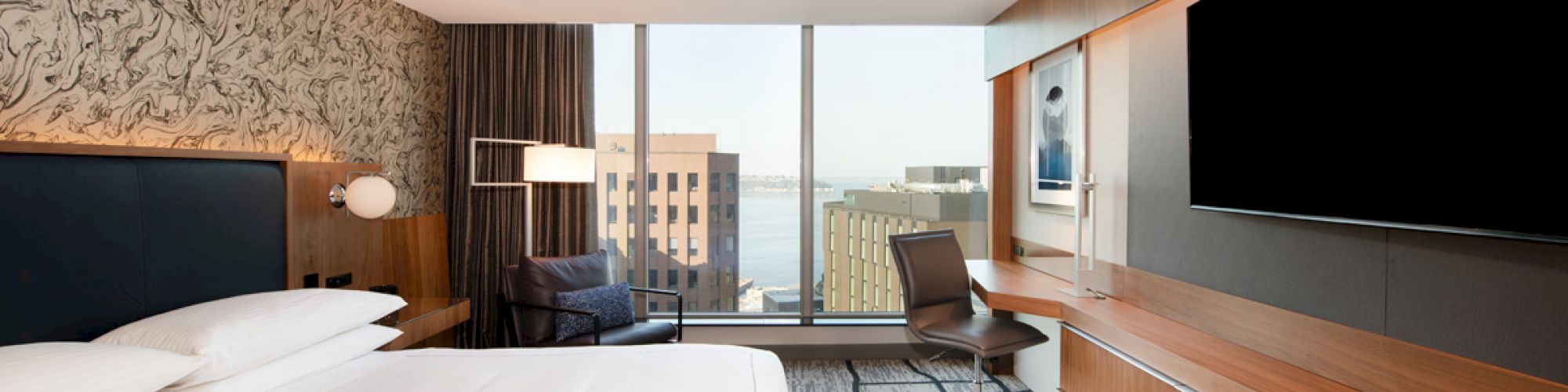 A modern hotel room with a large bed, wall-mounted TV, desk, chair, and city view through large windows.